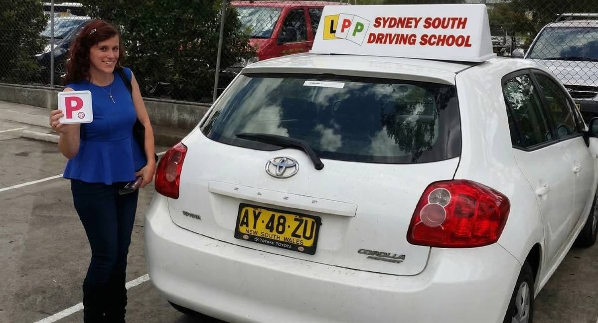 A student holding a red P plate beside a Sydney South Driving School dual-control Toyota Corolla