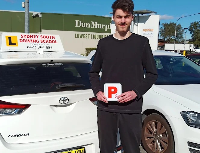 A Sydney South Driving School student holding a red P plate beside the school's white Toyota Corolla